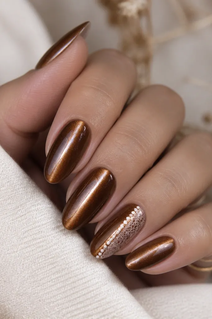 Five medium almond nails. Nutmeg brown cat eye stripe runs vertically. On the ring finger, there's a nude lace-like curve made of tiny connected dots and short lines along one side of the nail.