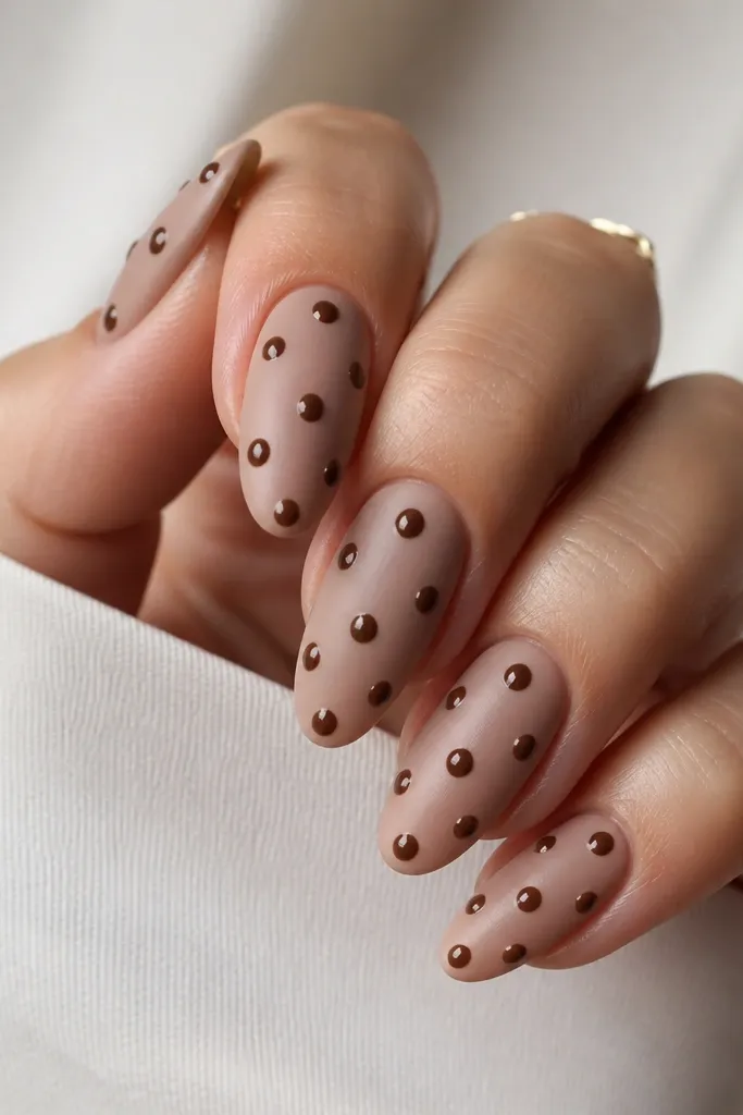 Nails with a matte nude base. Over the matte surface, glossy brown polka dots stand out with a shiny dome effect.