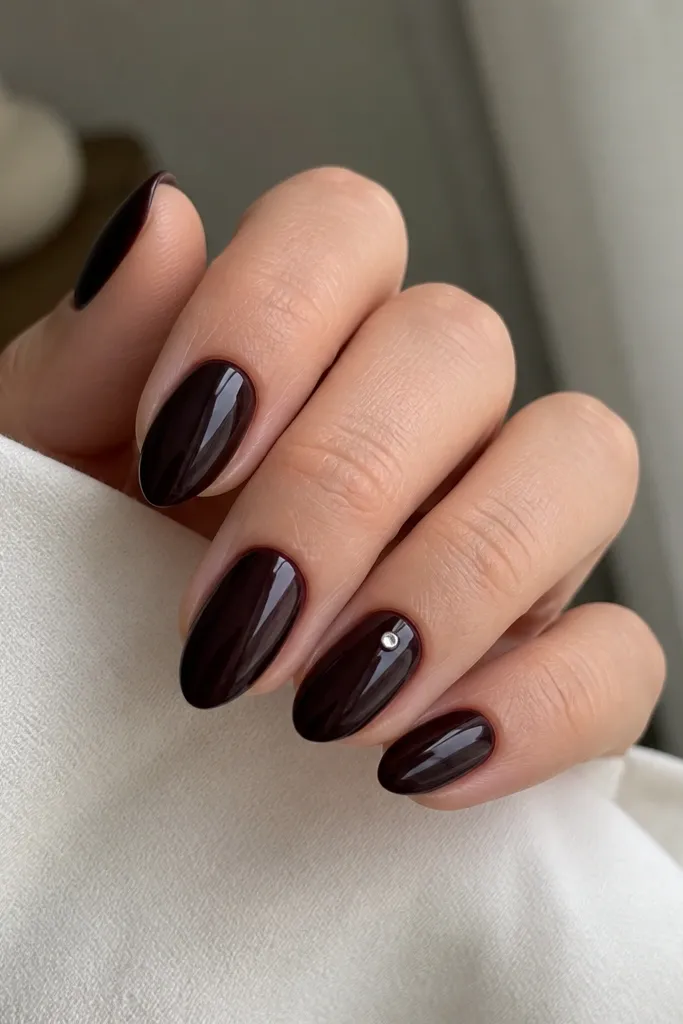 Short stiletto nails painted black cherry (dark red-black). On the ring finger, there's one tiny clear gem placed near the center of the nail, sitting on top of the gloss.