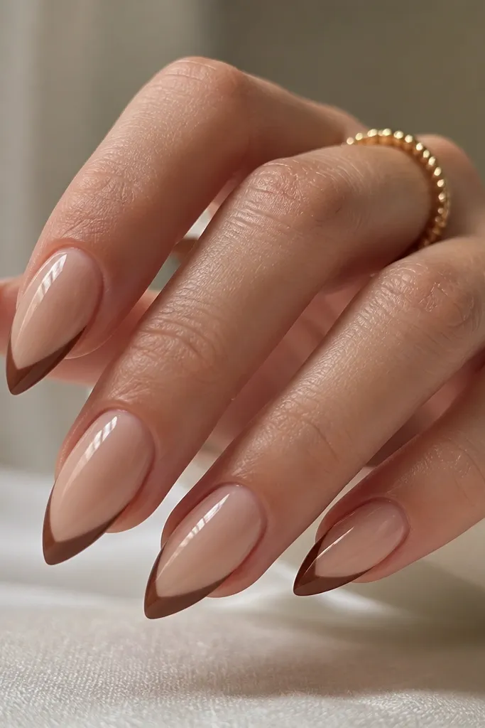 Short stiletto nails in a nude base. A thin chocolate-brown chevron sits on the free edge, pointing down toward the nail tip point. Glossy finish.