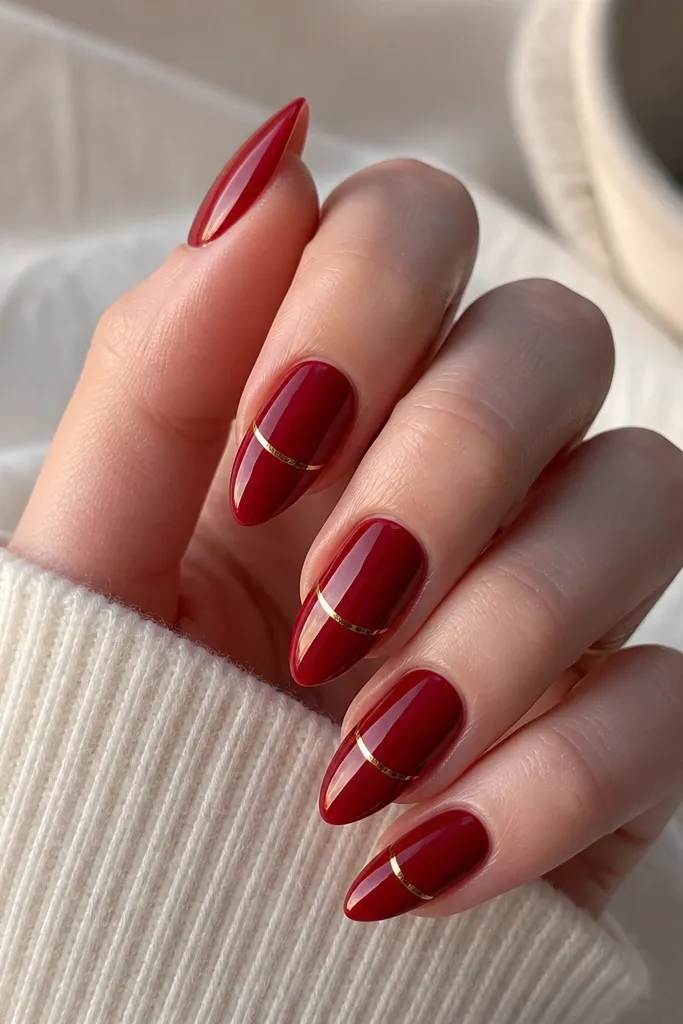Short stiletto nails painted deep red with a slightly velvety shine. A thin gold arc line curves along the free edge from one side to the other. Glossy top coat.
