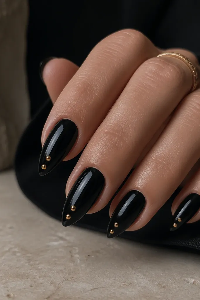 Short stiletto nails painted deep black. Two to three tiny gold studs sit along the free edge on each nail, clustered near the center and aligned with the taper. Glossy finish.