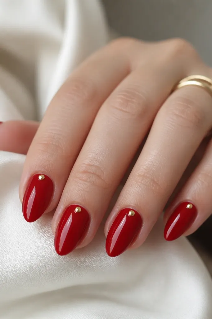 Short stiletto nails in opaque lipstick red. Each nail has one tiny gold foil dot placed right at the cuticle center, like a small jewel. Everything is glossy.
