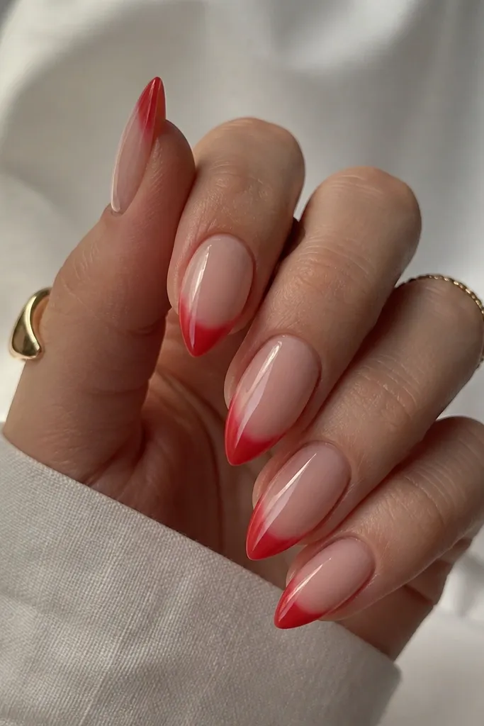 Short stiletto nails with a nude center and a red ombré tip. The red forms a French-like curved band at the top of the tip, fading into the point. Glossy finish.