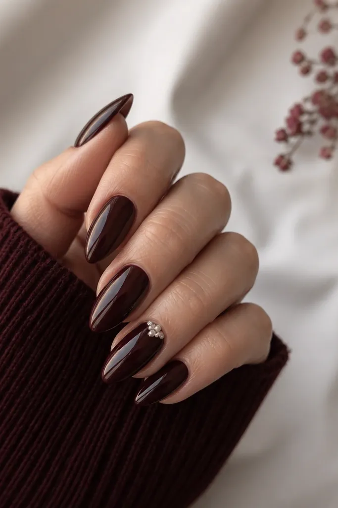 Short stiletto nails in oxblood glossy. One accent nail has a small cluster of tiny pearl-like beads near the cuticle, arranged in a tight group. Other nails are solid oxblood.