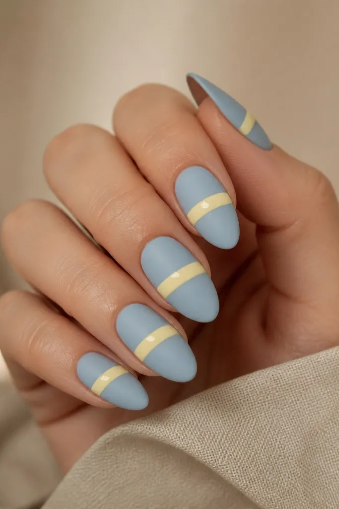Short squoval nails are matte powder blue across all nails. On each ring finger, there's a single glossy yellow stripe running vertically from cuticle to tip. The rest stays matte.