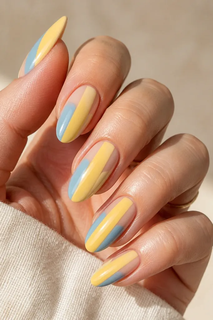 One nail per hand has a solid sheer nude base with a thick vertical yellow stripe. The same nail has a blue half-side panel on the cuticle side. Other nails are solid light blue.