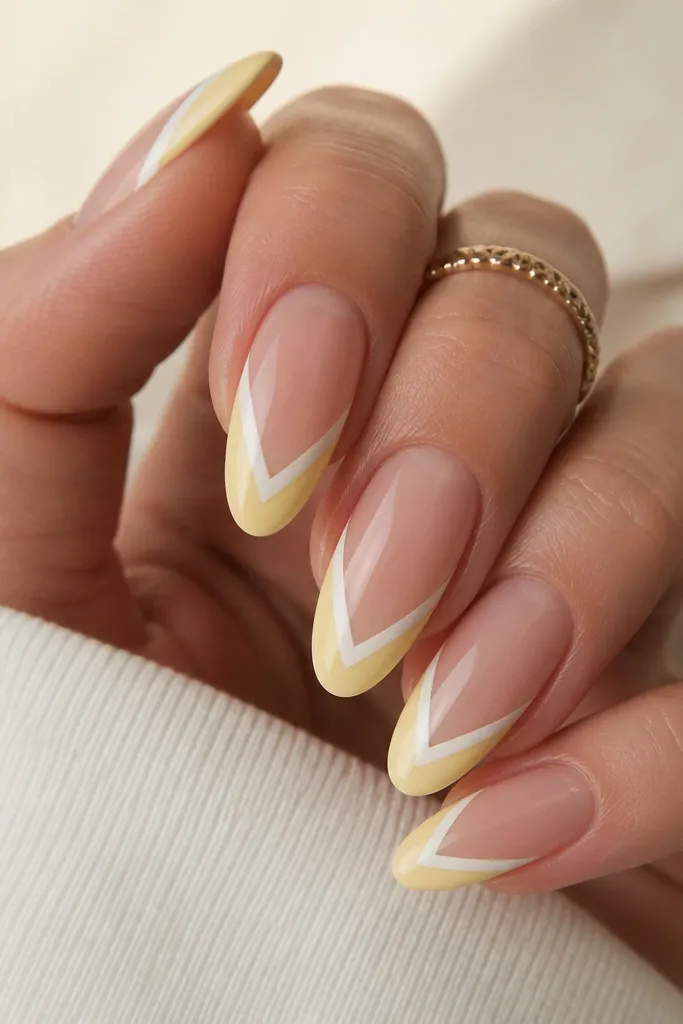 Almond nails with a creamy yellow French tip. The tip ends form a soft V shape in the center, with a thin white line tracing the V. Base is nude satin.