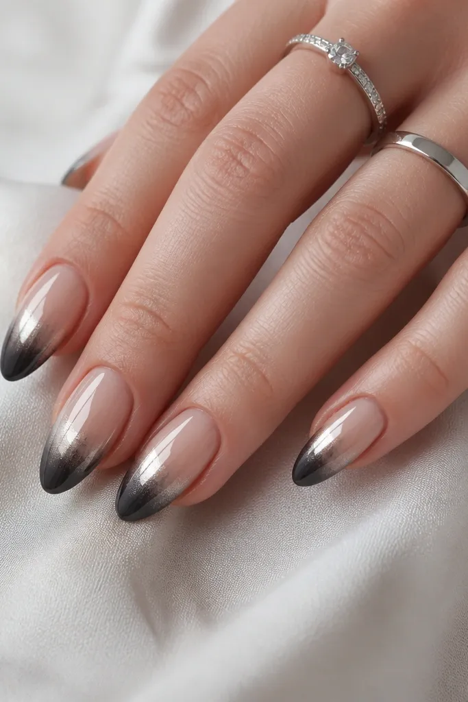 Medium almond nails. Base is sheer nude. Tips fade from deep charcoal-black at the edge upward into smoky gray. Silver chrome concentrates on the upper part of the fade, creating a reverse effect. High gloss.