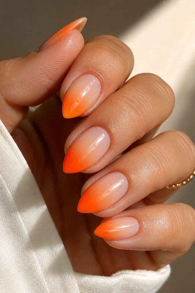 Short stiletto nails with a nude base fading into bright tangerine orange at the tips, glossy ombre look.