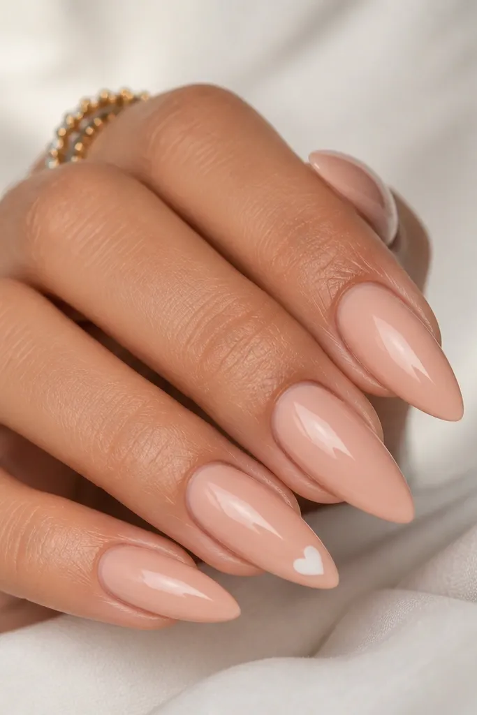 Short stiletto nails in soft peach nude; one accent nail has a tiny white heart near the tip, glossy finish.