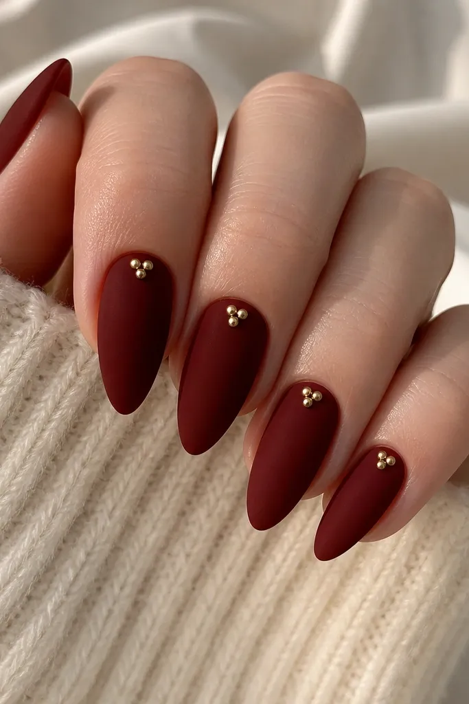 Short stiletto nails in deep red velvet matte; each nail has 3 tiny gold studs clustered near the cuticle center.