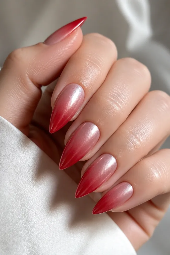 Short stiletto nails with a red ombré that fades into a softer, pearlized sheen; the whole nail looks glossy with a pearly glow.
