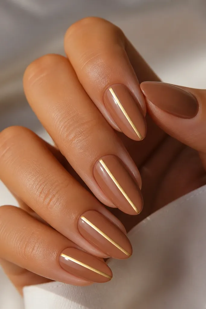 Four natural almond nails painted in a creamy light brown. A single bright gold stripe runs straight down the center of each nail with a glossy finish.