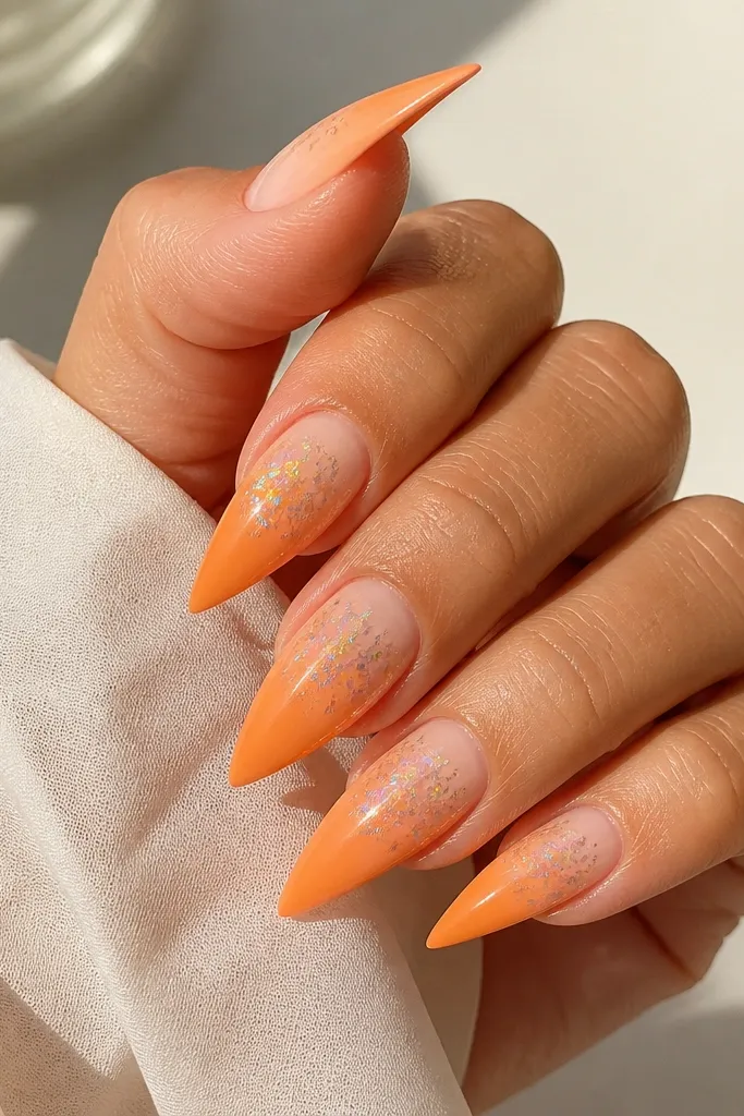 Stiletto nails with a tangerine ombre fading from pale peach at cuticle to bright orange at tips. Holo chrome is applied as small flicks across the top third, not covering the whole surface.