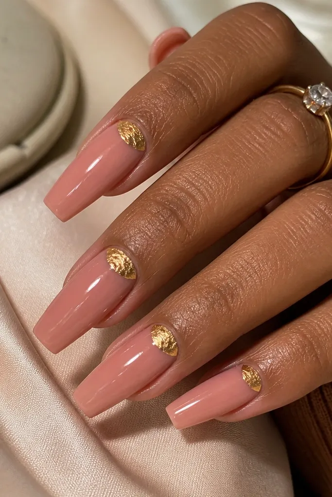 Glossy warm blush coffin nails with irregular gold foil half-moon shapes hugging the cuticle on three accent nails.