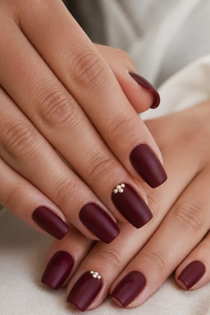 Short coffin nails in deep burgundy with a satin-leaning finish and tiny micro pearl clusters near the cuticle on accent nails.