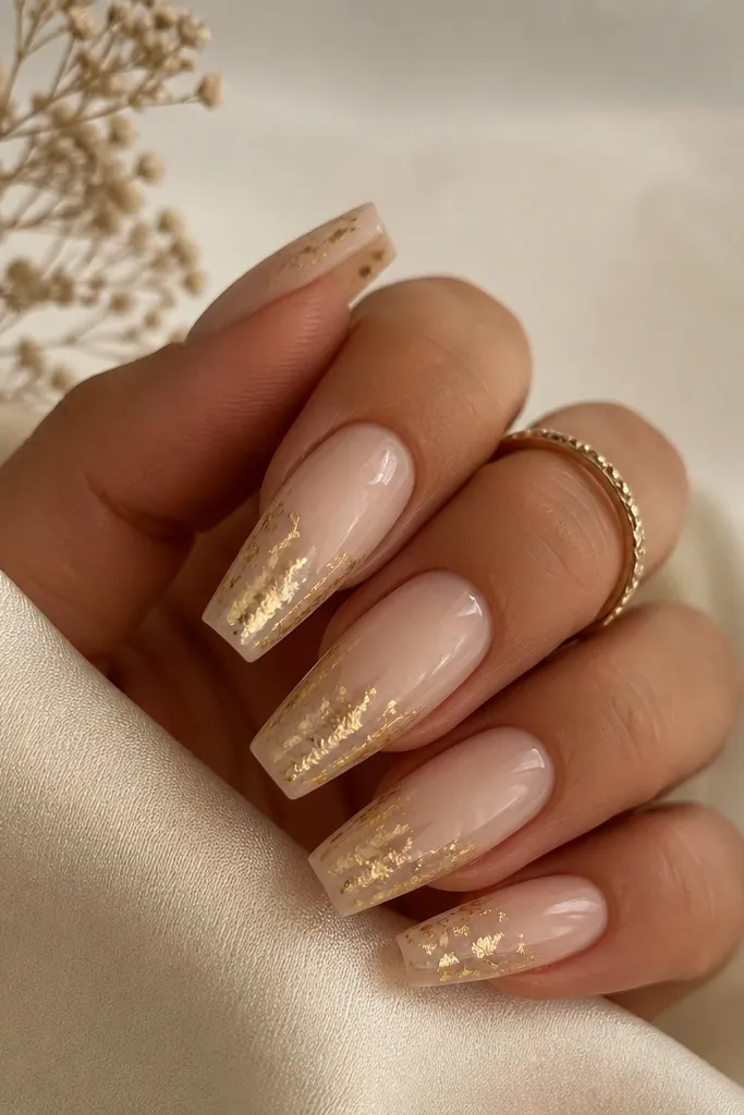 Champagne nude coffin nails with a warm milky base. Gold leaf is placed irregularly on the last third of each nail, concentrated near the tip and sealed under glossy topcoat.