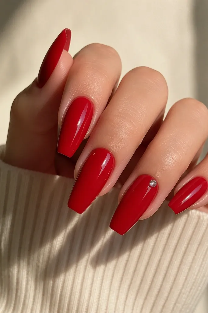 Glossy red coffin nails with one small crystal set near the cuticle on the ring finger, angled toward the side.
