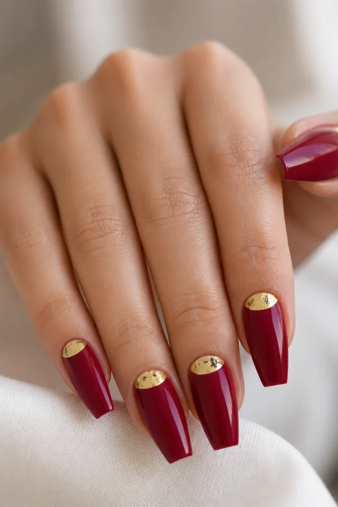 Coffin nails painted deep berry red. On each nail, there's a gold foil half-moon shape near the cuticle but not touching it, with the rest of the nail glossy and smooth.