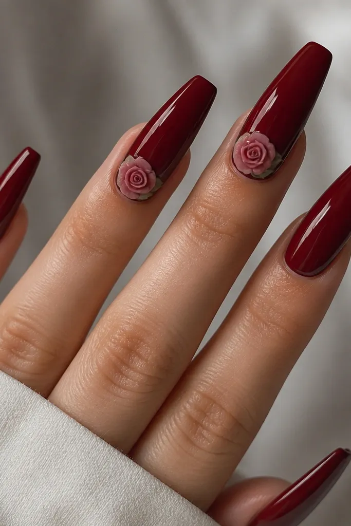 Five long coffin nails in a deep cherry red. Two nails have tiny 3D rose buds at the cuticle area, painted in muted rose-pink with glossy petals. The rest are solid cherry red with a high-gloss top coat.