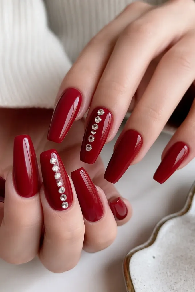 Cherry red coffin nails with a vertical ladder of small clear rhinestones on two nails. The rhinestones form a straight line from near the cuticle toward the center. The stones are glossy and reflective.