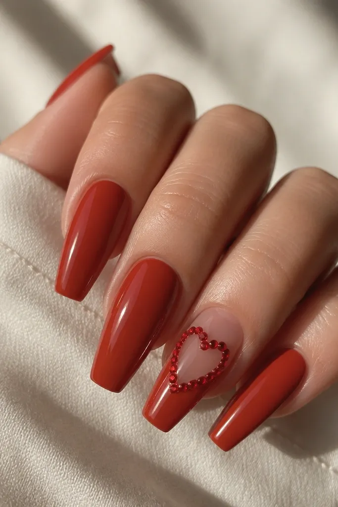 Warm red coffin nails with a negative space heart on one nail. The heart is outlined with small red crystals, and the center stays clear/glossy. The rest of the nails are solid warm red.