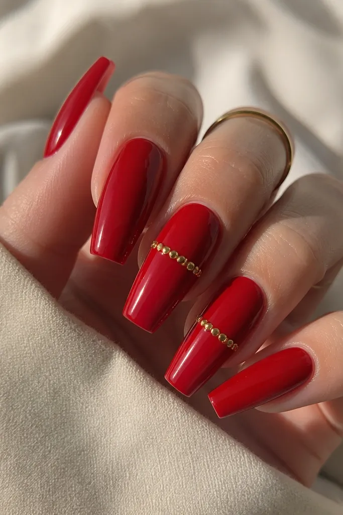 Warm red glossy coffin nails with a single horizontal row of tiny gold studs on the middle of two nails. The studs are evenly spaced and reflective. The rest of the nails are solid warm red.