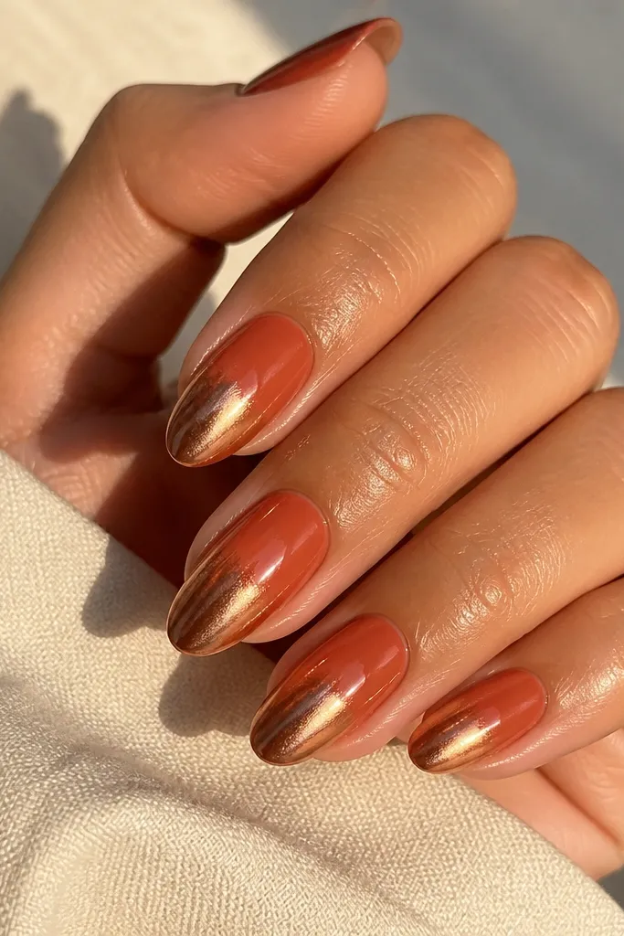 Medium almond nails with a terracotta orange base fading into rose gold chrome at the tips. The chrome is warm and reflective, with a smooth transition and a glossy coat that catches golden light.