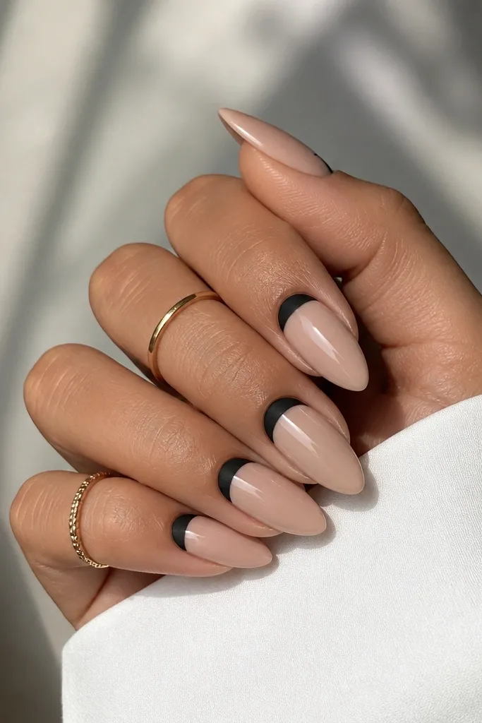 Short stiletto nails with nude base and a black satin reverse French near the cuticle. The matte black area sits in a smooth half-moon shape at the base of each nail, with the rest nude and glossy.