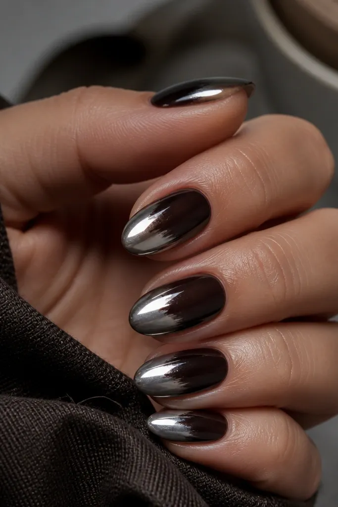 Short almond nails with a deep espresso brown almost-black ombre that fades into cool steel chrome at the tips. The chrome is reflective and slightly gray, with strong mirror highlights.