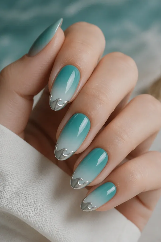 Medium almond nails with a teal sea-glass ombre that fades into a pale aqua near the tip. Silver chrome forms a wave-shaped tip detail, following the free edge like a tiny crest.