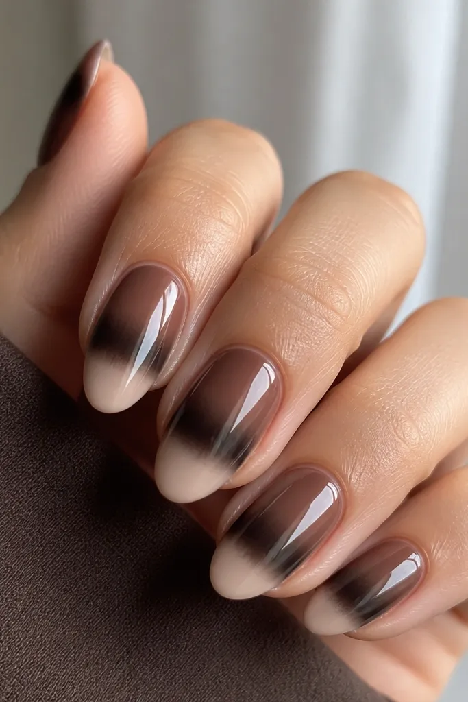 Short almond nails with a mocha brown ombre fading to a nude beige tip. Black chrome creates a smoky French band that looks blurred at the edges.