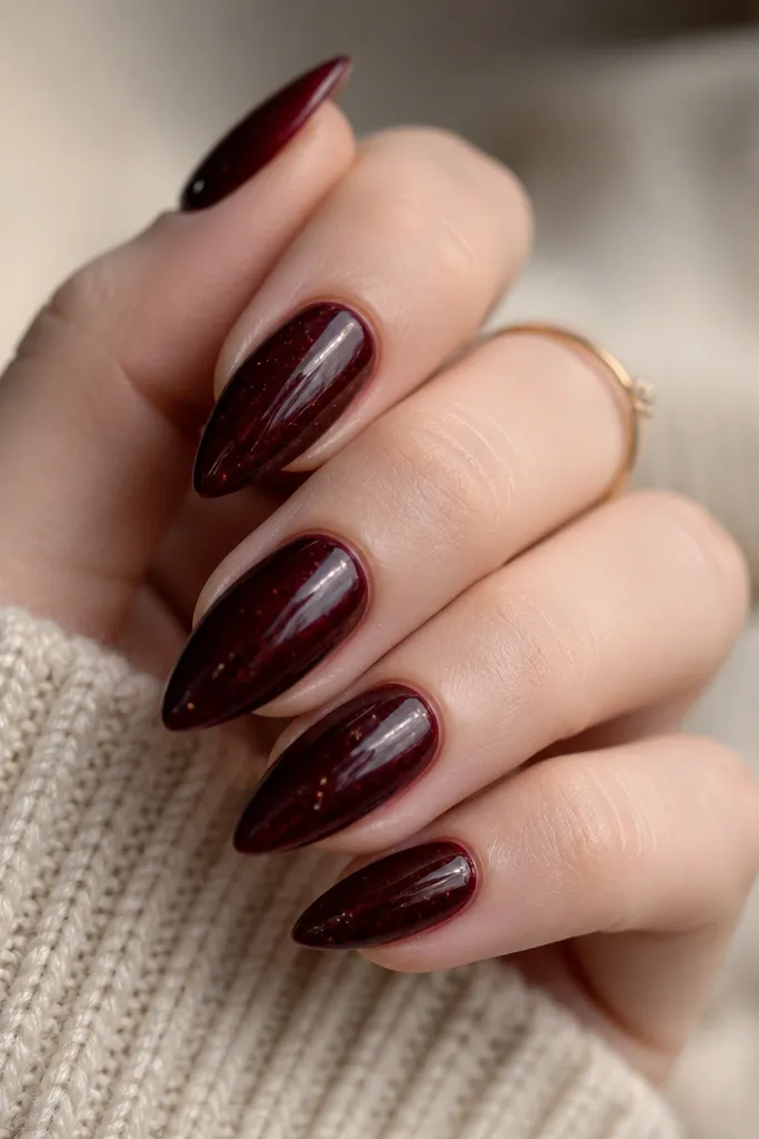 Short stiletto nails in a dark black cherry red with subtle gold flecks suspended in the polish. The nails have a glossy topcoat and the flecks are scattered lightly.