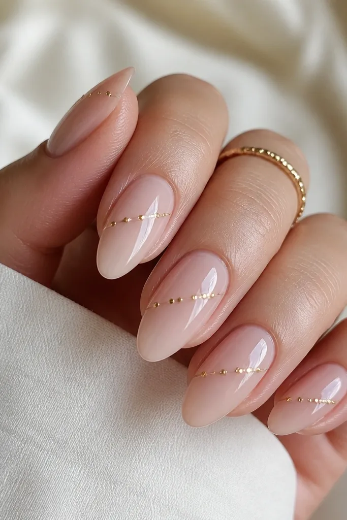 Nude pink almond nails with a glassy finish. Tiny gold micro beads are embedded in a diagonal line across each nail, suspended in the gel.