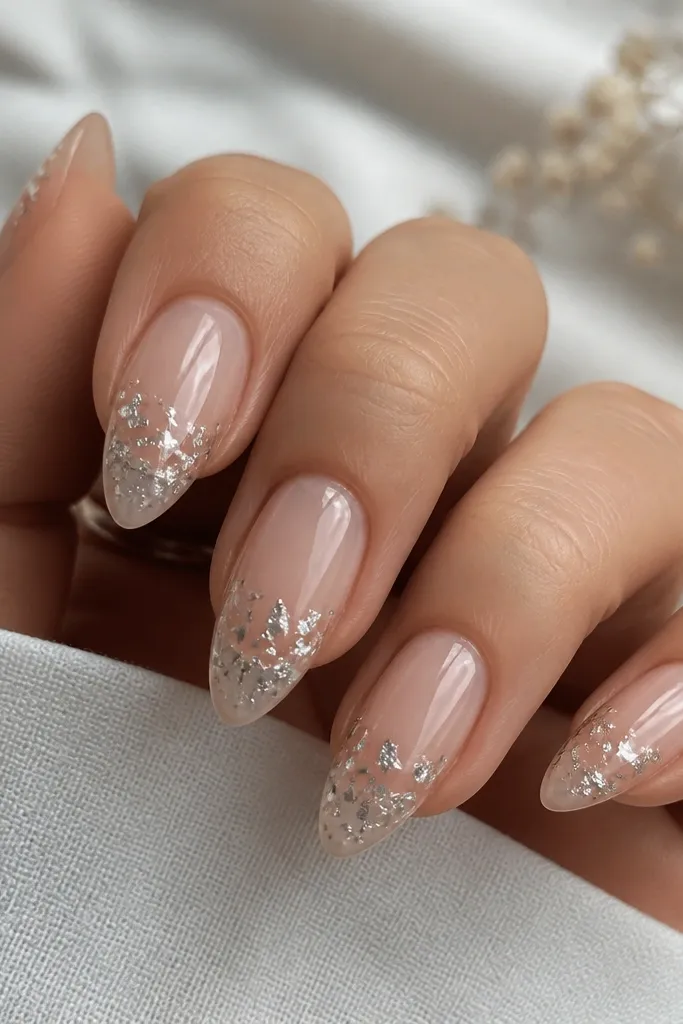 Short stiletto nails with a clear nude look, glossy. Tiny silver foil flakes sit mostly in the lower half near the free edge, like confetti.