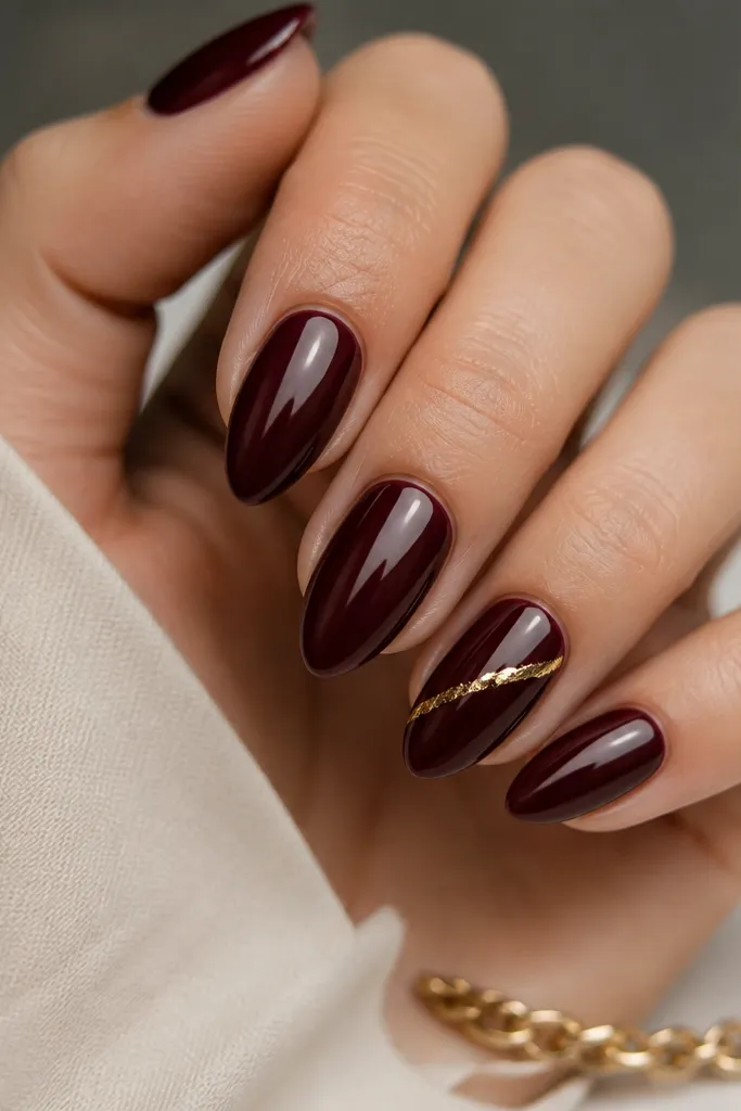 Short stiletto nails painted deep burgundy, mirror glossy. One nail has a narrow diagonal gold foil stripe across the middle.