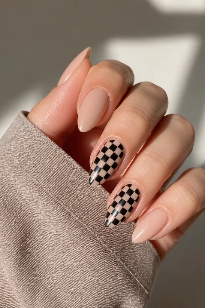 Short stiletto nails: three nails are solid nude glossy. Two nails have a tiny black-and-nude checkerboard pattern across the center with crisp square edges.