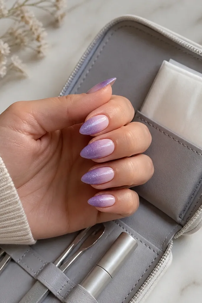 Short stiletto nails with lavender shimmer that fades from deeper purple at the tips to a pale lavender near the cuticle. A folded pack of lint-free wipes sits in a small side pocket.