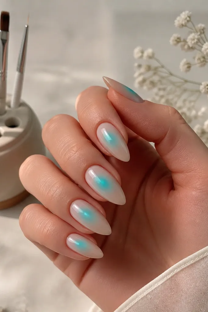 Short stiletto nails with a nude base. A turquoise glow effect sits around the center like an aura, with soft edges and a faint white halo. A small gel brush holder is shown with brushes separated.