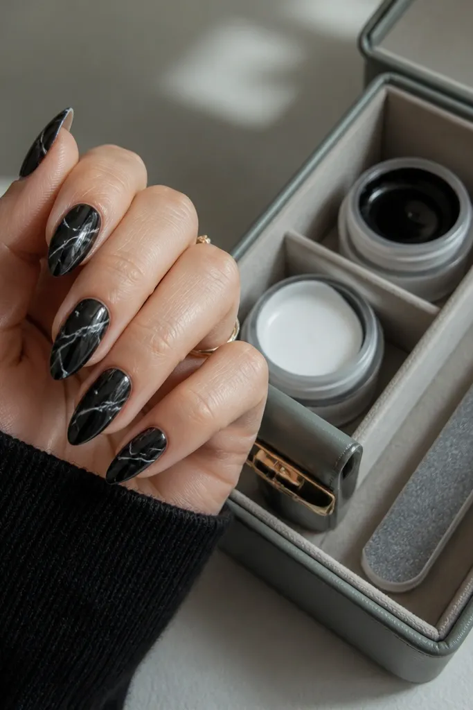 Short stiletto nails with deep black base and white marble veins flowing diagonally. Beside the nails is a storage organizer with separate compartments for liner gel, top coat, and a file.