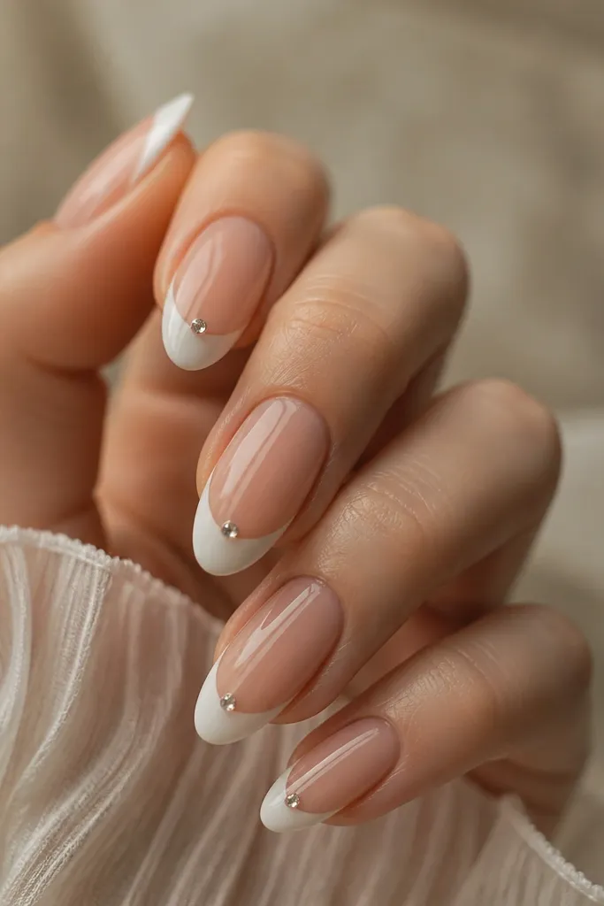 Short nails with nude base and thin white French tips. One small clear rhinestone sits exactly in the center of the smile line on each nail. Glossy top coat around the stone.