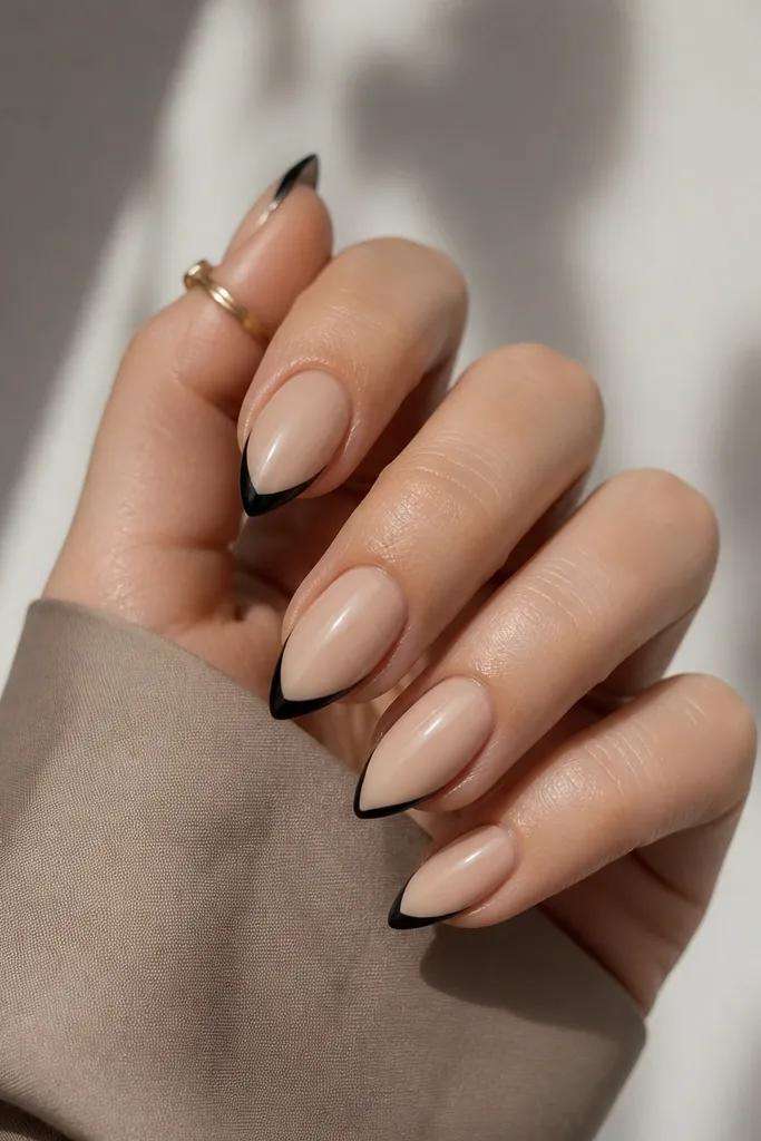 Short stiletto nails with a nude beige base. The french tips are tiny and crisp in matte-black or satin-black, forming a narrow smile line and a sharp tapered point. The nails look modern and minimal.