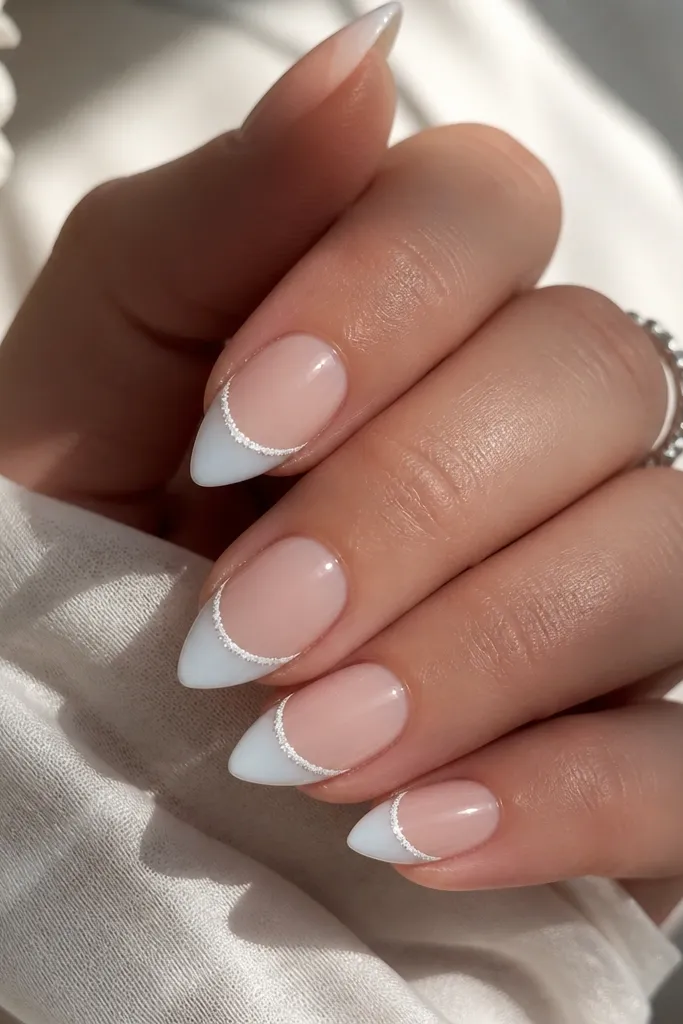 Short stiletto nails with a sheer nude base and icy milky white french tips. Along the smile curve, there's a thin line of fine silver micro-glitter. The glitter line is straight and narrow.