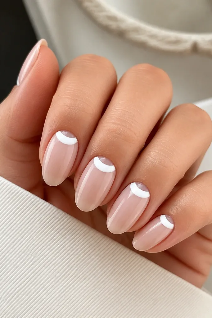 Short almond nails. The base is sheer nude. At the cuticle, there's a clear negative-space half-moon, and a white crescent overlay wraps around it, glossy and clean.