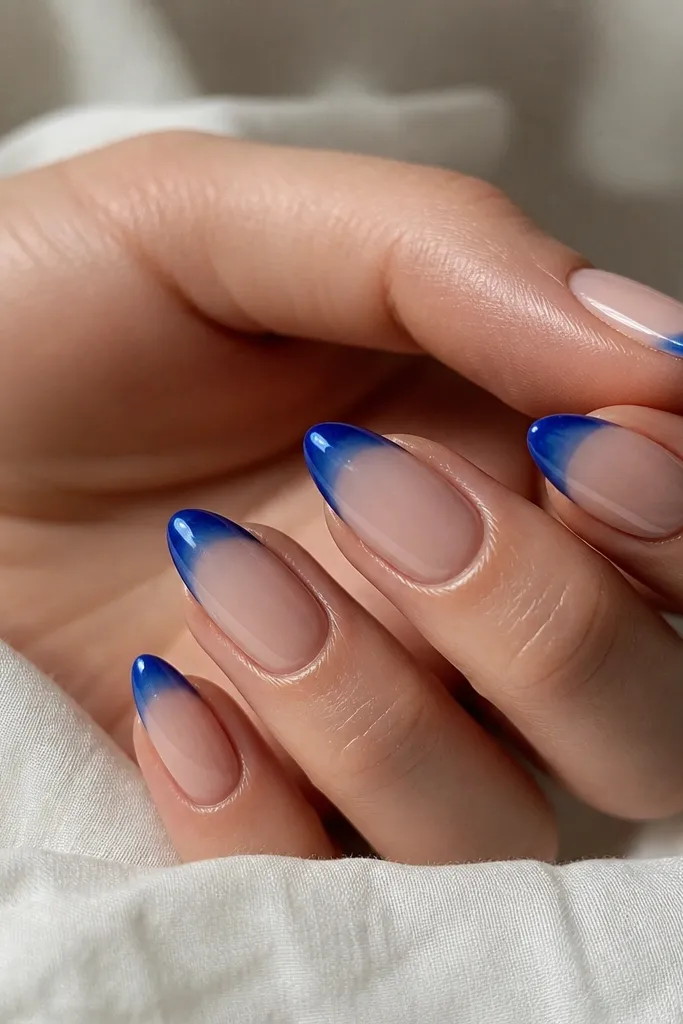 Short stiletto nails with a sheer nude base. The tips are cobalt blue in a glossy, glassy gradient that looks like liquid. Cuticles are clean and edges are sharp.