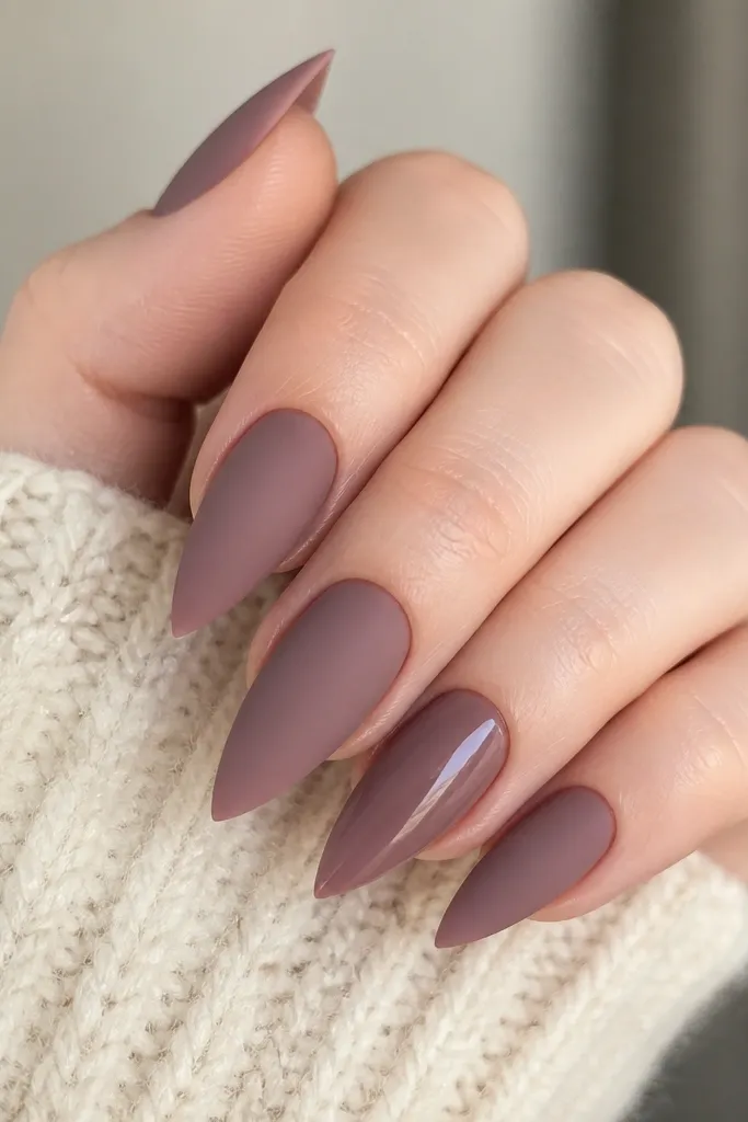 Short stiletto nails in smoky mauve. Most nails are matte, but one accent nail has a glossy mauve with a soft shine. Cuticles are neat and the taper is sharp.