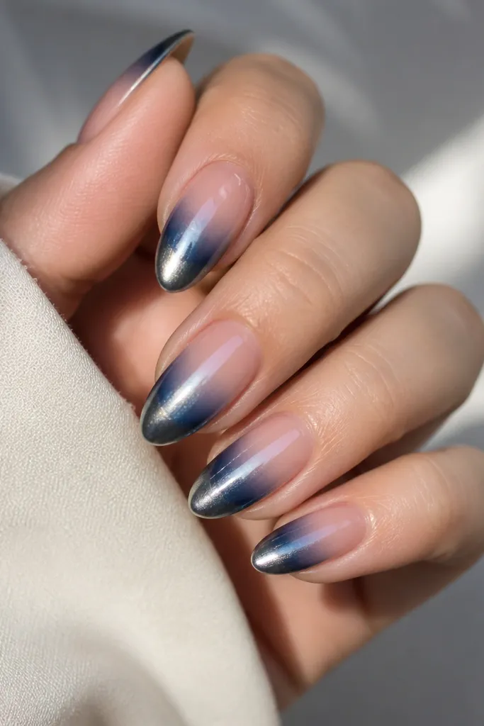 Almond nails with sheer pink at the cuticle fading into soft navy at the tips. Steel chrome is applied near the tip and fades upward, like a glow around the navy edge.