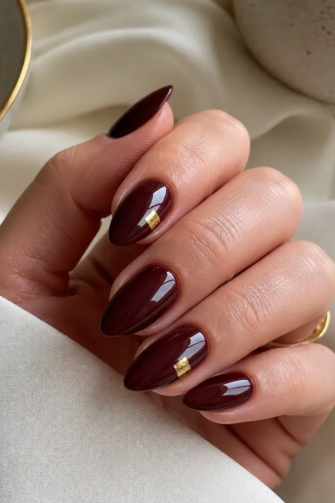 Short stiletto nails in deep oxblood red with a high-gloss finish. On two accent nails, there's a small gold foil square placed near the outer corner of the nail, not across the whole tip.