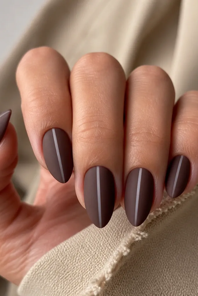 Short stiletto nails painted espresso brown in a satin-matte finish. Each nail has a thin vertical clear strip down the center, creating negative space. The cuticles are clean and the clear strip is perfectly straight.